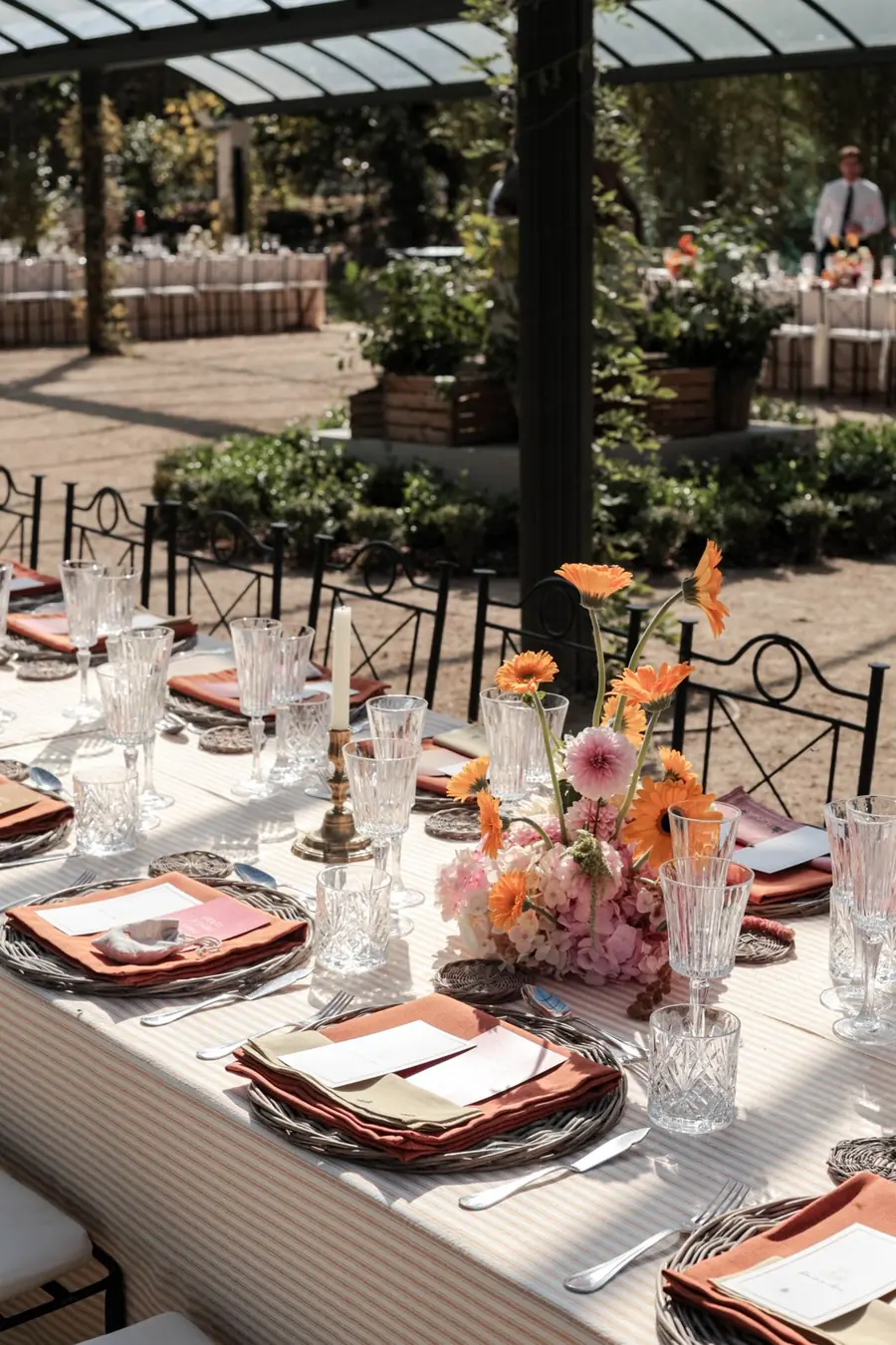 Mesa larga bajo una pérgola, con flores naranjas y bajoplatos de mimbre